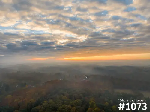 Luchtfoto van herfstbos bij Beek-Ubbergen met mist tussen de bomen en een oranje gloed onder een wolkenlucht