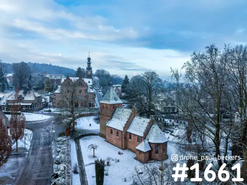 Luchtfoto van een bakstenen kapel met torentje in de sneeuw in Beek-Ubbergen, met kerktoren achter kale bomen