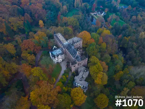 Luchtfoto van een kasteel met leistenen daken in Beek-Ubbergen, omringd door bos in herfstkleuren van geel, oranje en rood