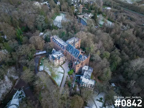 Luchtfoto van een kasteel van rode baksteen met torentjes in het bos bij Beek-Ubbergen, sneeuwresten op het gazon