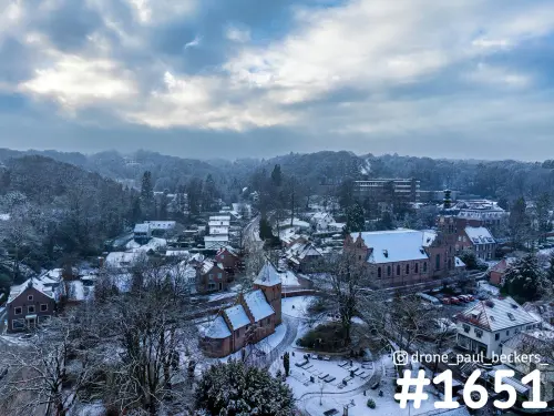 Luchtfoto van Beek-Ubbergen in de sneeuw, met een bakstenen kerk en besneeuwde daken tegen beboste heuvels