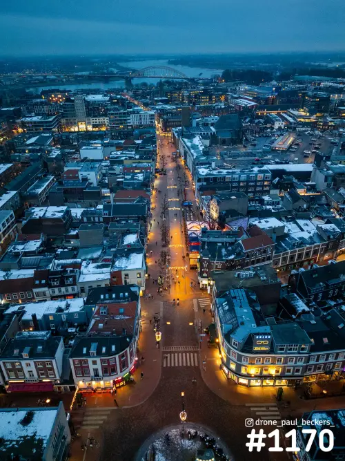Luchtfoto van het centrum van Nijmegen in de schemering, met besneeuwde daken en een verlichte straat naar de Waalbrug
