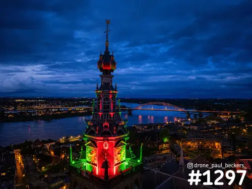 Luchtfoto van het centrum van Nijmegen in de avond, met een rood en groen verlichte toren en de Waalbrug over de Waal
