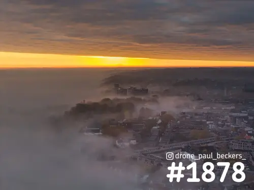 Luchtfoto van het centrum van Nijmegen in dichte mist bij zonsopkomst, met een oranje horizonlijn boven het Valkhof gebied