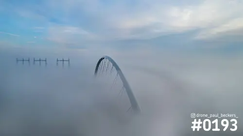 Luchtfoto van de boog van De Oversteek in Nijmegen die boven een dik mistdek uitsteekt, met hoogspanningsmasten links in de verte