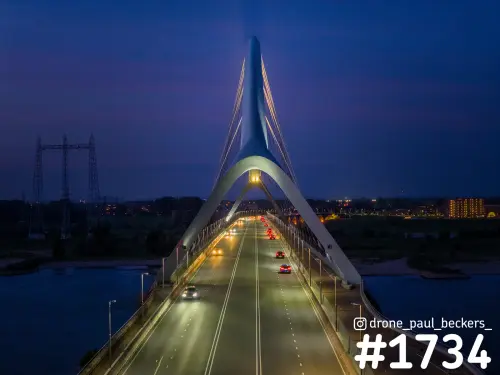 Luchtfoto van De Oversteek in Nijmegen in de avondschemering, met brugboog, verlichte rijbaan en auto's richting Lent