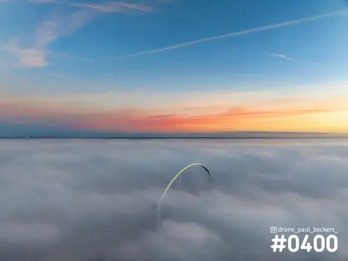 Luchtfoto van de boog van De Oversteek bij Nijmegen die boven een dik mistdek uitsteekt, met een oranje gloed aan de horizon