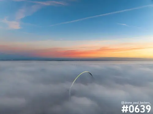 Luchtfoto van de boog van De Oversteek in Nijmegen die boven een dik mistdek uitsteekt, met een oranje en roze gloed aan de horizon