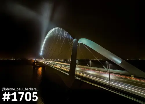 Luchtfoto van de Oversteek in Nijmegen 's nachts, met lichtstrepen van auto's op het wegdek en de verlichte boog in de mist