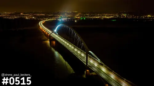 Luchtfoto van de verlichte boogbrug De Oversteek in Nijmegen bij nacht, met de stadslichten aan de horizon