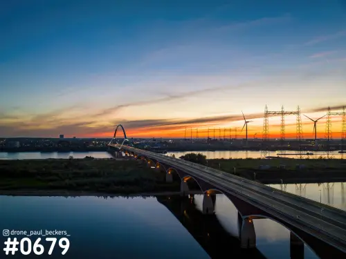 Luchtfoto van de brug De Oversteek over de Waal bij Nijmegen na zonsondergang, met oranje gloed en windturbines aan de horizon