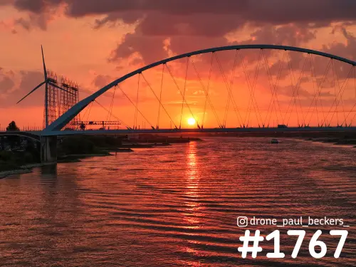 Luchtfoto van de boogbrug De Oversteek over de Waal in Nijmegen bij zonsondergang, met rood gloeiende lucht en een windmolen links