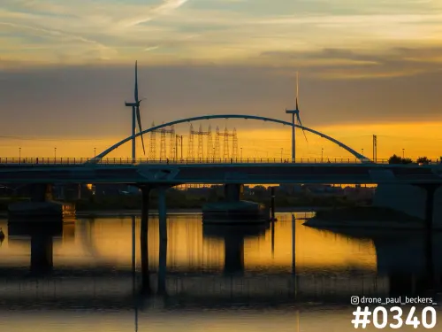 De Oversteek in Nijmegen bij zonsondergang, met de boogbrug, twee windturbines en hoogspanningsmasten achter het spiegelende water