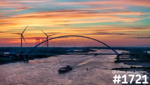 Luchtfoto van De Oversteek over de Waal bij Nijmegen na zonsondergang, met vrachtschip, windmolens en oranje wolkenlucht