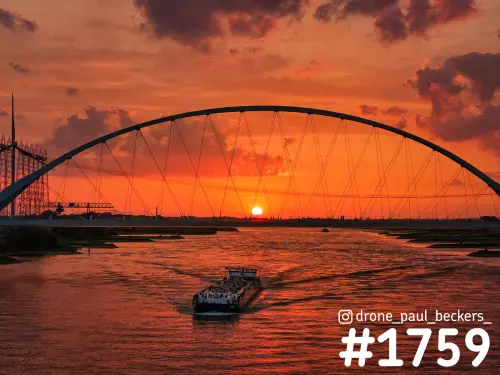 Luchtfoto van De Oversteek over de Waal bij Nijmegen, met een binnenvaartschip en de zon onder de boog in een oranjerode lucht