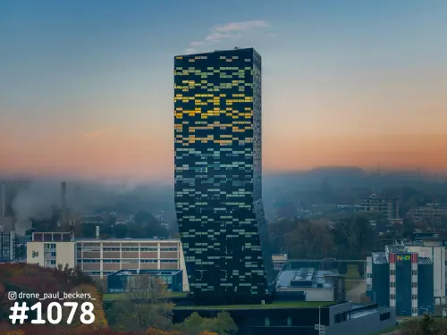Luchtfoto van een hoge glazen toren bij de Goffert in Nijmegen met verlichte ramen, oranje ochtendgloed en herfstbomen