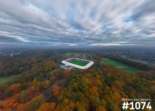 Luchtfoto van stadion De Goffert in Nijmegen, omgeven door herfstbos met oranje bomen onder een brede wolkenlucht