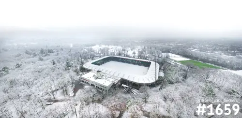Luchtfoto van stadion De Goffert in Nijmegen onder een laag sneeuw, met besneeuwde bomen en een groen veld ernaast