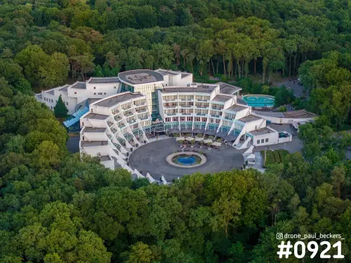 Luchtfoto van een wit gebogen gebouw met fontein op rond plein midden in het bos, Goffert omgeving Nijmegen, zomeravond