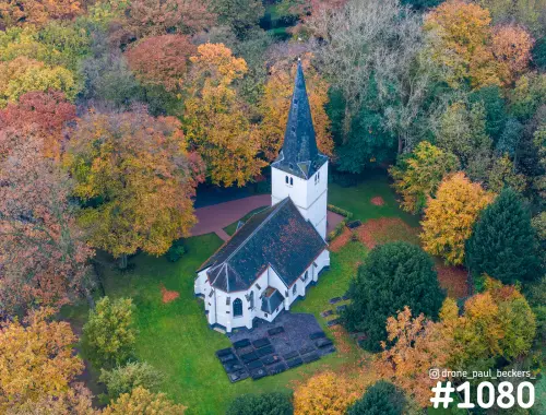 Luchtfoto van een wit kerkje met donker dak en spitse toren, omringd door herfstbomen bij de Goffert in Nijmegen