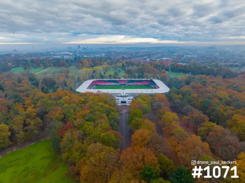 Luchtfoto van het Goffertstadion in Nijmegen, omringd door herfstbomen in oranje en geel onder een bewolkte lucht