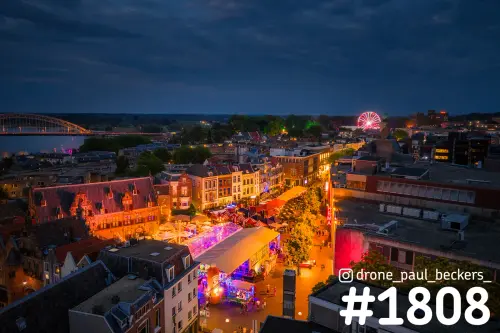 Luchtfoto van de Grote Markt in Nijmegen in de avond, met verlichte feesttenten, een reuzenrad en de Waalbrug links