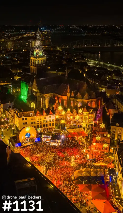 Luchtfoto van de Grote Markt in Nijmegen bij avond, met een drukke menigte, verlichte kramen en de Stevenskerk erachter