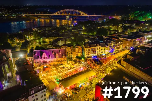 Luchtfoto van de Grote Markt in Nijmegen 's avonds met verlichte feesttenten en de Waalbrug op de achtergrond