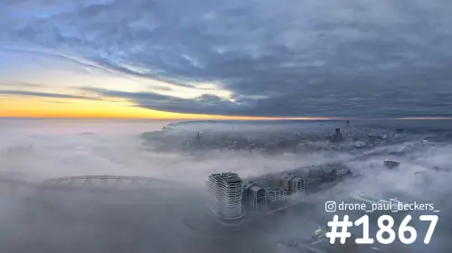 Luchtfoto van de Handelskade in Nijmegen in dichte mist, met de stad op de achtergrond en een oranje gloed onder zware wolken