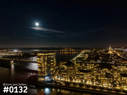 Luchtfoto van de Handelskade in Nijmegen bij nacht, met maan boven de Waal, de Waalbrug en de Stevenskerk in de verte