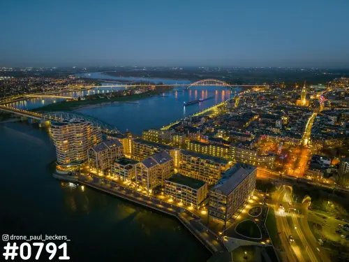 Luchtfoto van de Handelskade in Nijmegen in de blauwe uurtje-avond, met verlichte woongebouwen aan de Waal en de Waalbrug