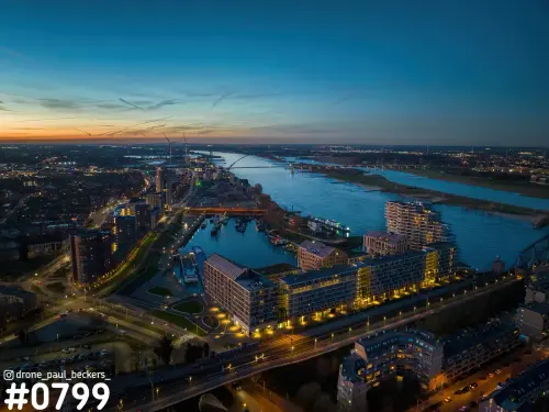 Luchtfoto van de Handelskade in Nijmegen bij schemering, met verlichte appartementen, de jachthaven en de Waal met De Oversteek