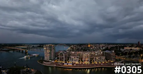 Luchtfoto van de Handelskade in Nijmegen in de schemering, met verlichte appartementen aan de Waal en een dreigend wolkendek