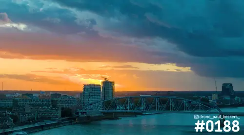 Luchtfoto van de Waal bij de Handelskade in Nijmegen met stalen brug, hoogbouw en een oranje avondlucht onder donkere wolken