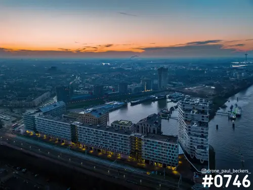 Luchtfoto van de Handelskade in Nijmegen bij schemering, met woonblokken langs het spoor en de Waal onder oranje lucht