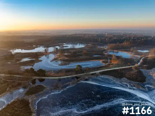 Luchtfoto van de Hatertse Vennen bij zonsopkomst, met deels bevroren vennen, slingerend zandpad en heide in oranje licht