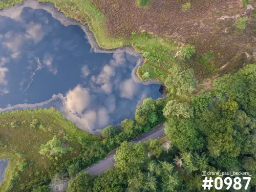 Luchtfoto van een ven in de Hatertse Vennen met wolken in het water, paarse heide en een smalle asfaltweg door het groen