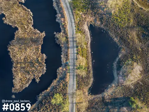 Luchtfoto recht van boven van een zandpad tussen twee vennen in de Hatertse Vennen, met lange boomschaduwen over het pad