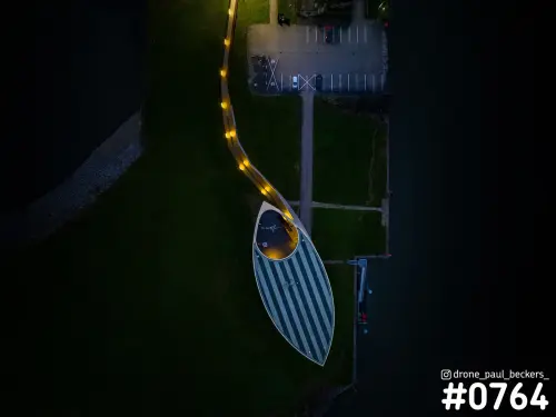 Luchtfoto van een bladvormig gebouw met gestreept dak en verlicht pad aan het water, avond in het havengebied van Nijmegen