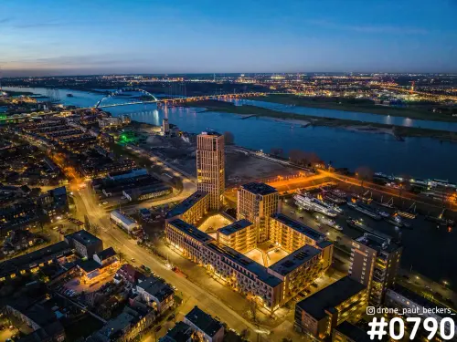 Luchtfoto van het Havenkwartier in Nijmegen in het blauwe uur, met verlichte woongebouwen, de Waal en De Oversteek op de achtergrond