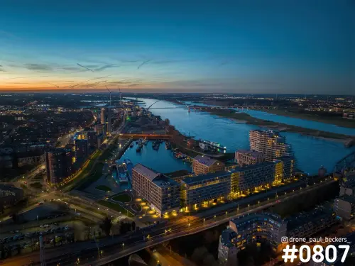 Luchtfoto van het Havenkwartier in Nijmegen bij schemering, met verlichte woonblokken aan de haven en de rivier de Waal
