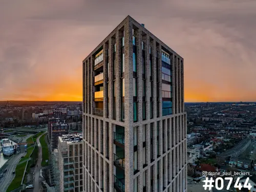 Luchtfoto van een hoge bakstenen woontoren in het Havenkwartier met oranje gloed aan de horizon en een schip in de haven