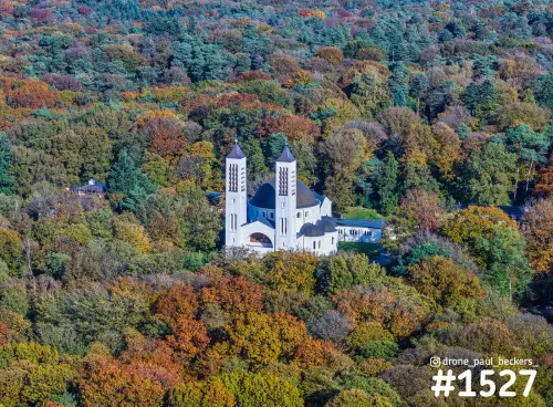 Luchtfoto van de witte kerk met twee torens op de Heilig Landstichting, omringd door herfstbos in geel, oranje en groen