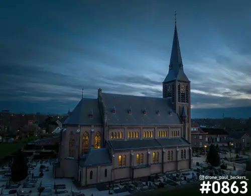 Luchtfoto van de verlichte kerk in Weurt in de avondschemering, met kerkhof en gele gloed achter de glas-in-loodramen