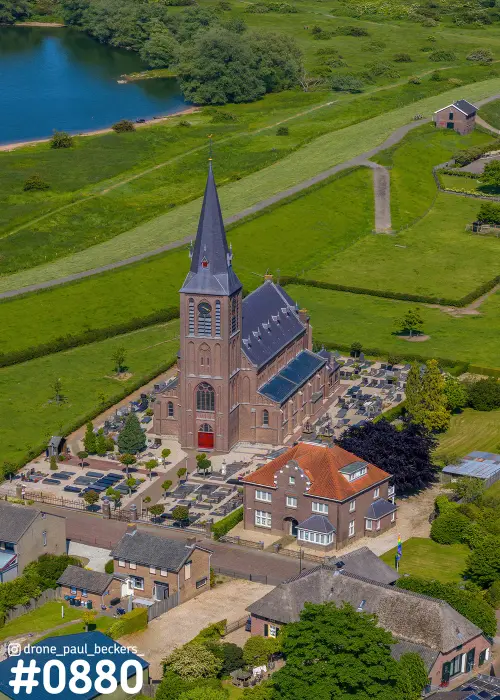 Luchtfoto van de kerk van Weurt bij Nijmegen met bakstenen toren, begraafplaats en zonnepanelen op het dak, omgeven door groene dijkweiden