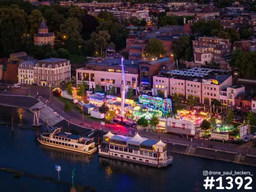 Luchtfoto van de kermis langs de Waal in Nijmegen in de schemering, met verlichte attracties en twee afgemeerde boten