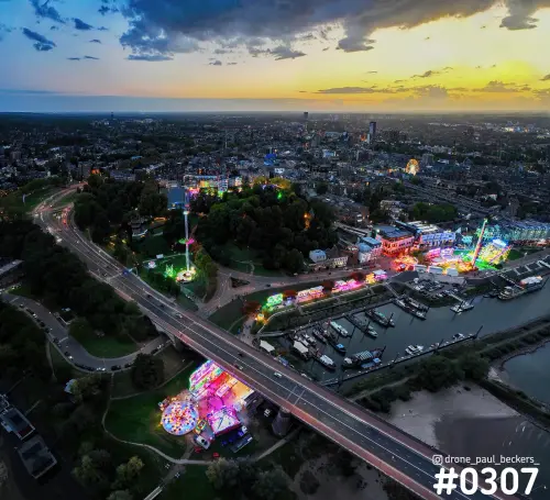 Luchtfoto van de kermis aan de Waalkade in Nijmegen bij zonsondergang, met verlichte attracties naast de Waal en de Lindenberghaven