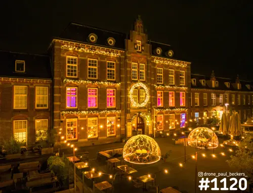 Luchtfoto van een verlicht baksteengebouw met kerstverlichting en lichtgevende iglotenten op het terras in de avond