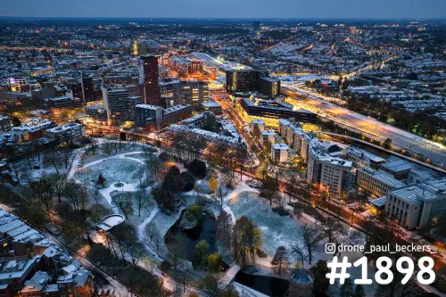 Luchtfoto van het Kronenburgerpark in Nijmegen in de schemering met sneeuw, met verlichte hoogbouw en het stationsgebied erachter