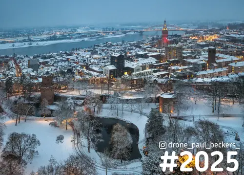 Luchtfoto van besneeuwd Nijmegen in de schemering, met Kronenburgerpark, verlichte Stevenskerk en de Waal erachter